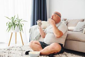 Eats and does exercises. Funny overweight man in casual clothes is indoors at home photo