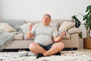 Eats and does exercises. Funny overweight man in casual clothes is indoors at home photo