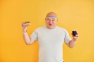 con comida rápida. el hombre gracioso con sobrepeso en la corbata de cabeza deportiva está contra el fondo amarillo foto