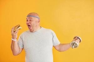 Holds hamburger and dumbbell. Funny overweight man in sportive head tie is against yellow background photo