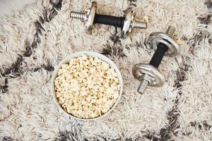 Top view of popcorn in white plate and dumbbells that lying down on the floor photo