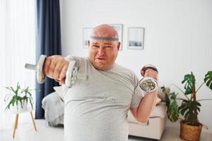 utiliza mancuernas. un hombre gracioso con sobrepeso vestido con ropa informal está en el interior de su casa foto