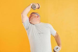 Exercises by using dumbbells. Funny overweight man in sportive head tie is against yellow background photo