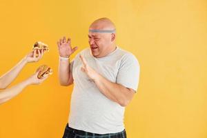 Hands with hamburgers temptates guy. Funny overweight man in sportive head tie is against yellow background photo