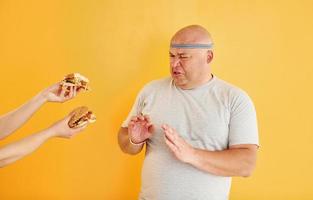 Hands with hamburgers temptates guy. Funny overweight man in sportive head tie is against yellow background photo