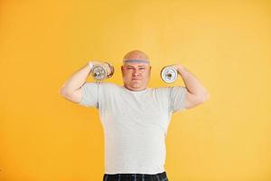 Exercises by using dumbbells. Funny overweight man in sportive head tie is against yellow background photo