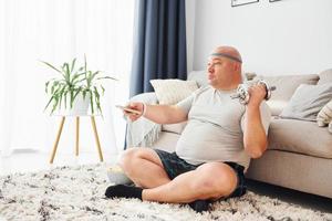 Eats and does exercises. Funny overweight man in casual clothes is indoors at home photo