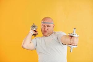 Exercises by using dumbbells. Funny overweight man in sportive head tie is against yellow background photo