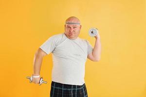 Exercises by using dumbbells. Funny overweight man in sportive head tie is against yellow background photo