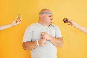 manzana o galleta. el hombre gracioso con sobrepeso en la corbata de cabeza deportiva está contra el fondo amarillo foto
