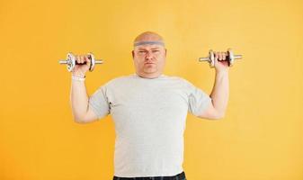 Exercises by using dumbbells. Funny overweight man in sportive head tie is against yellow background photo