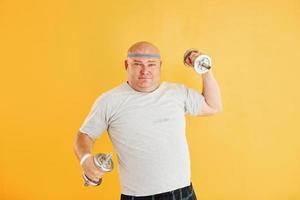 Exercises by using dumbbells. Funny overweight man in sportive head tie is against yellow background photo