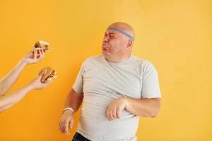 Hands with hamburgers temptates guy. Funny overweight man in sportive head tie is against yellow background photo