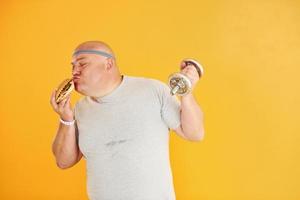 Holds hamburger and dumbbell. Funny overweight man in sportive head tie is against yellow background photo