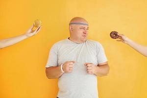 Apple or cookie. Funny overweight man in sportive head tie is against yellow background photo