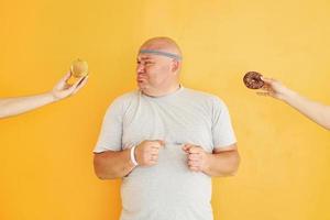 Apple or cookie. Funny overweight man in sportive head tie is against yellow background photo