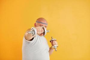 Exercises by using dumbbells. Funny overweight man in sportive head tie is against yellow background photo