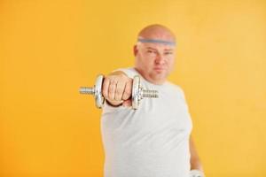 Exercises by using dumbbells. Funny overweight man in sportive head tie is against yellow background photo