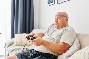 Plays video game. Funny overweight man in casual clothes is indoors at home photo