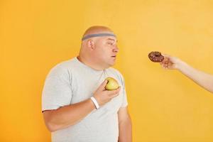 manzana o galleta. el hombre gracioso con sobrepeso en la corbata de cabeza deportiva está contra el fondo amarillo foto