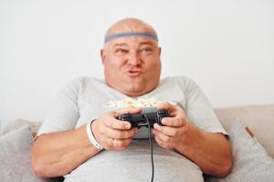 Plays video game. Funny overweight man in casual clothes is indoors at home photo