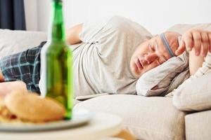 Hamburgers and beer. Man lying on the sofa. Funny overweight man in casual clothes is indoors at home photo