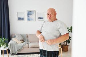 medir la cintura. un hombre gracioso con sobrepeso vestido con ropa informal está en el interior de su casa foto