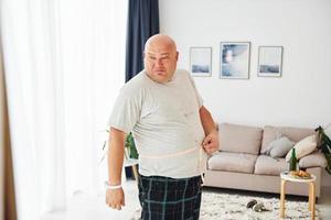 medir la cintura. un hombre gracioso con sobrepeso vestido con ropa informal está en el interior de su casa foto