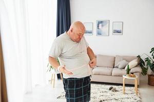 Measuring waist. Funny overweight man in casual clothes is indoors at home photo