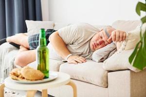 hamburguesas y cerveza. hombre acostado en el sofá. un hombre gracioso con sobrepeso vestido con ropa informal está en el interior de su casa foto