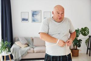 medir la cintura. un hombre gracioso con sobrepeso vestido con ropa informal está en el interior de su casa foto