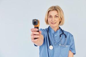 With infrared thermometer. Middle-aged professional female doctor in uniform and with stethoscope photo
