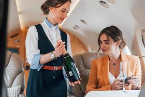 Stewardess with bottle of alcohol. Young passenger woman in yellow clothes is flying in the plane photo