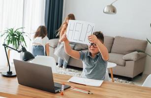 Boy holding paper with help word. Refuses to take a lesson. Kids having fun in the domestic room at daytime together photo