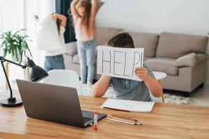 Boy holding paper with help word. Refuses to take a lesson. Kids having fun in the domestic room at daytime together photo