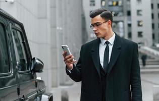 Using phone. Businessman in black suit and tie is outdoors in the city photo