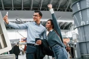 usando el teléfono para hacer selfie. pareja joven está en el aeropuerto juntos foto