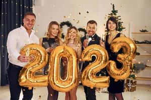 Holding balloons with 2023 number. Group of people have a new year party indoors together photo