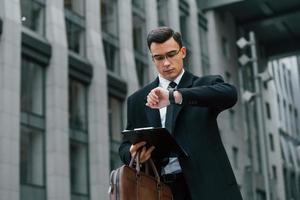 Looking at clock. Businessman in black suit and tie is outdoors in the city photo