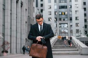 Looking at clock. Businessman in black suit and tie is outdoors in the city photo