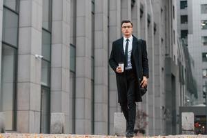 With cup of drink. Businessman in black suit and tie is outdoors in the city photo