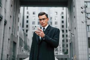 teléfono en la mano. hombre de negocios con traje negro y corbata está al aire libre en la ciudad foto