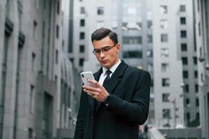 Phone in hand. Businessman in black suit and tie is outdoors in the city photo