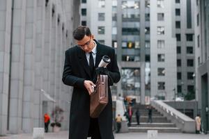 poner el teléfono en la bolsa. hombre de negocios con traje negro y corbata está al aire libre en la ciudad foto