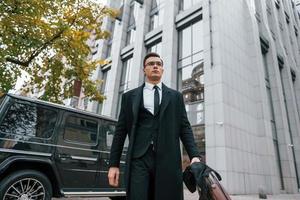 Standing against black car. Businessman in black suit and tie is outdoors in the city photo