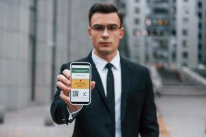 Holding phone with vaccination certificate. Businessman in black suit and tie is outdoors in the city photo