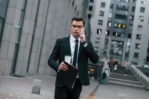 Walking and talking by phone. Businessman in black suit and tie is outdoors in the city photo