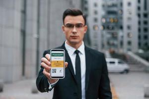 Holding phone with vaccination certificate. Businessman in black suit and tie is outdoors in the city photo