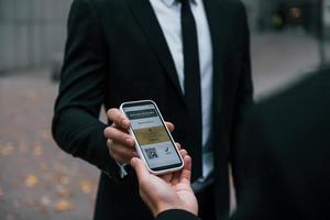 Holding phone with vaccination certificate. Businessman in black suit and tie is outdoors in the city photo