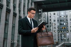 With notepad in hands. Businessman in black suit and tie is outdoors in the city photo